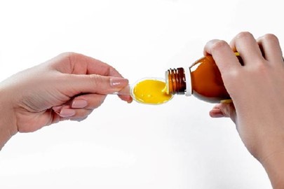 Hands pouring liquid medication from a brown bottle onto a spoon.