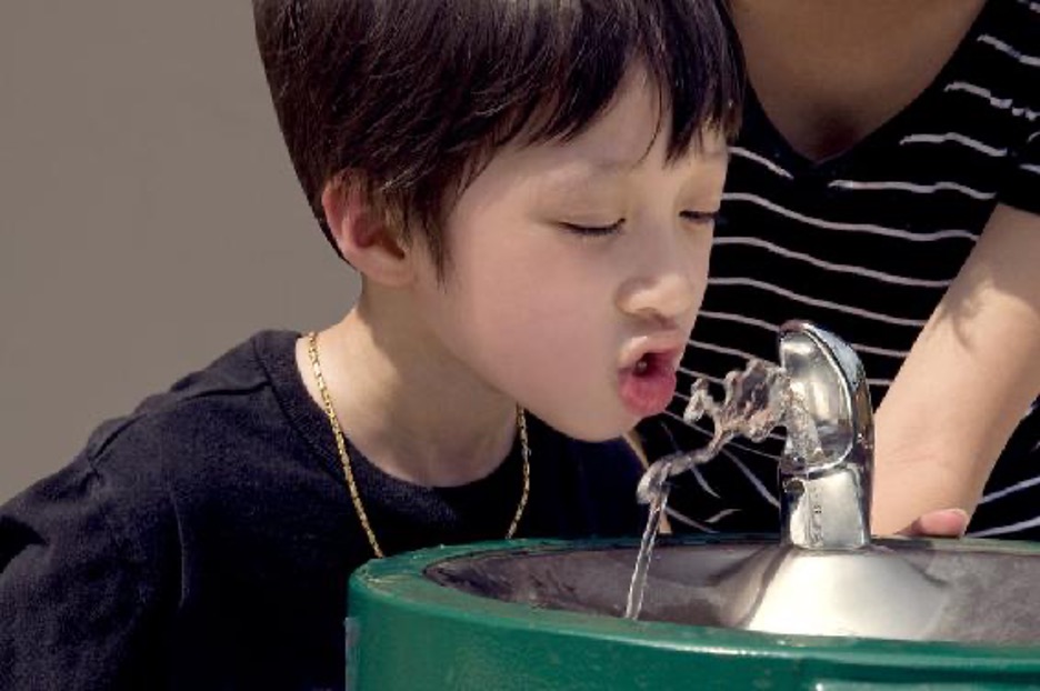A young boy stands behind a fountain taking a drink of water.