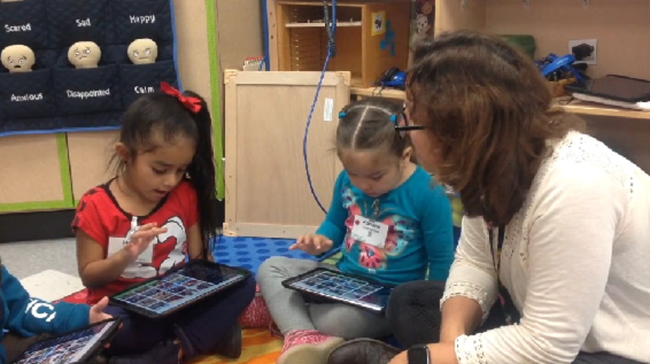 Two young girls sit on chairs and play on iPads under the supervision of an adult.