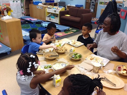 A group of toddlers sits around a table with plates of food in front of them.