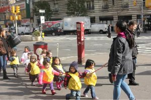 A line of toddlers wear bright yellow vests and walk in pairs while holding a rope between them.