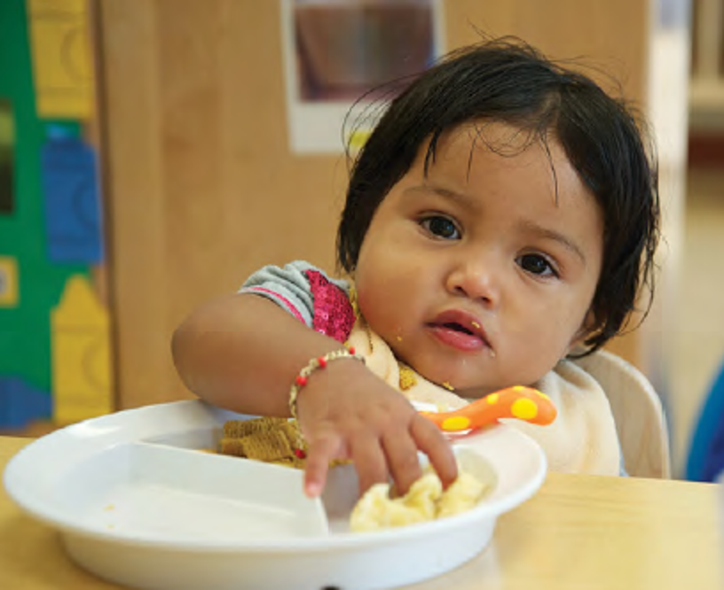 A little girl reaches into a bowl to grab solid food.