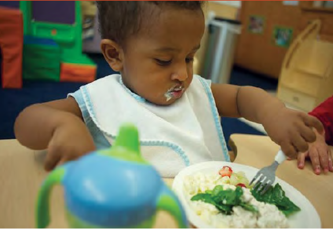 A toddler sitting in a high chair has a plate of food in front of them and is self-feeding.