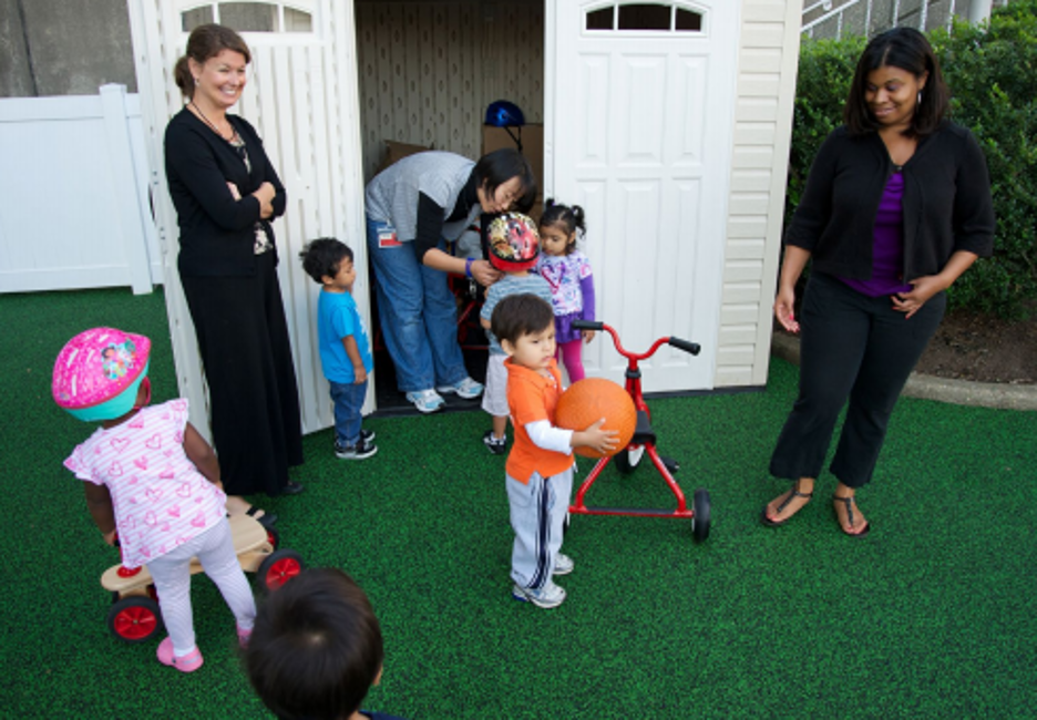 Children play outside on turf with tricycles under the supervision of adults.