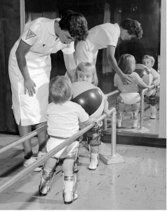 Two children with polio undergoing physical therapy; there is a ball between them and they are walking with the assistance of bars.