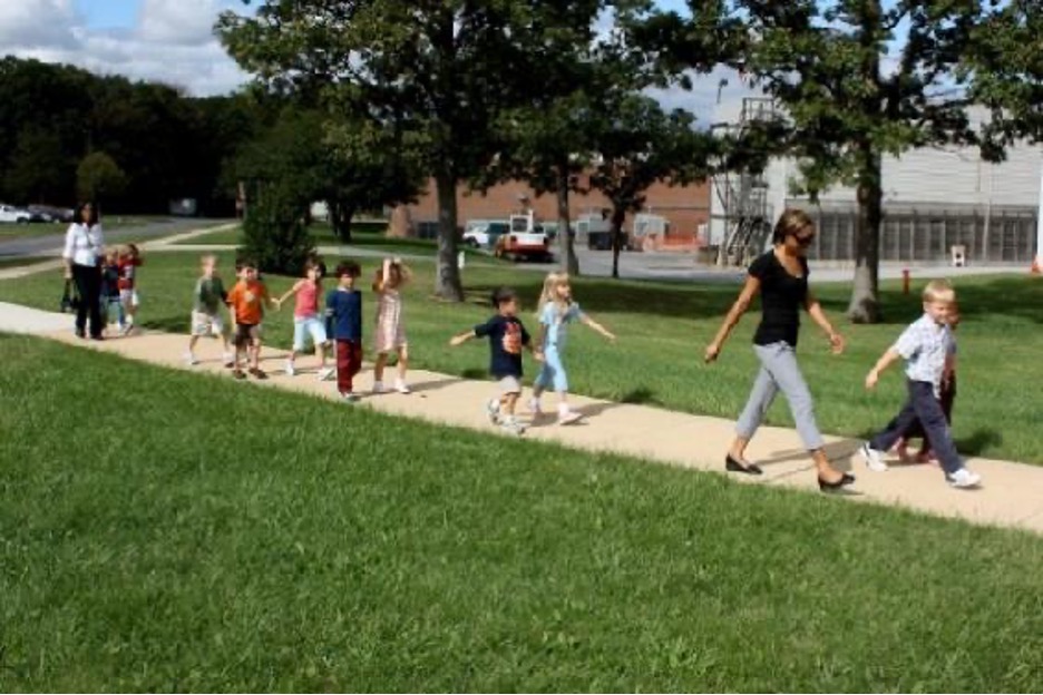 Two adults walk with a group of children in a line between them; the children hold hands in pairs.