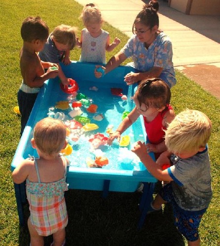 A group of young children stand around a water table and play with toys within it.