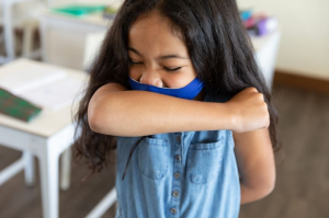 A young girl wearing a mask holds her arm in front of her mouth as she sneezes.