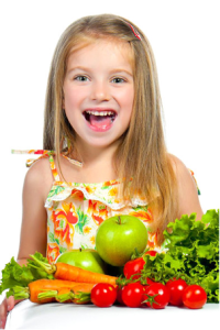 A smiling girl has a pile of fruits and vegetables on the table in front of her.