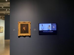 a photograph taken inside an art gallery showing a dark grey wall that has a framed painting and a video monitor side-by-side. The painting is framed in a gold-colored frame and is of a bored looking young girl in a dark dress and hat. The video monitor shows the same painting on the far right side of the screen while two artists communicate with one another using American Sign Language on the main portion of the screen. There is closed captioning in the bottom section of the screen.