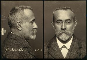 Two black and white photographs of the same size set side by side. The image is of a white man visible from the shoulders up with a neutral expression. On the left side image, the man is visible in profile, and on the right, he is facing the camera head-on. The man has short greying hair, a beard and a moustache. He is wearing a suit jacket with a collared white undershirt. The left image shows a three-pronged device used to keep the subject's head in one place. The device is thin and small, making contact with no more than two inches of the figure's head.