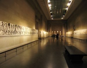 A photograph of the interior of a museum space. The room is dimply it, with the lighting focused on long rows of decorative marble carvings lining each wall. At the far end of this long room, five statues on blocks can barely be seen. The silhouette of a person walking towards these statues appears close to the centre of the image. A black bench is located at the bottom right of the photograph.