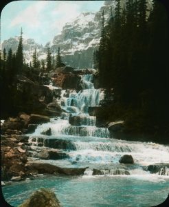 A photograph of a rocky waterfall. This multi-level waterfall is flanked by tall evergreen trees starts at a rocky elevation in the top third section of the image and terminates at a pool of blue-green water in the foreground. A mountain range capped with snow is visible in the distance.