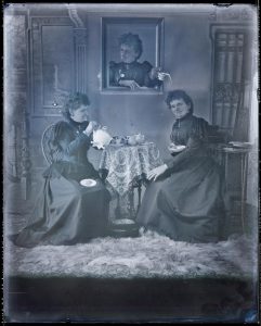 A multiple exposure black and white photograph showing the same woman appearing 3 times. She wears a long dark dress. Two versions of the woman sit and have tea at a table while another appears in a picture frame on the wall, pouring tea onto the head of one of the woman sitting at the table.