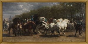 A rectangular oil painting in a thin light-coloured wood frame. A busy scene comprised of multiple figures comprising of horses and humans takes up the central section of the entire painting. The horses are of various colours, white brown and black. The horses are ridden or led by men of varying descriptions. The men seem to be predominantly white, however, some skin tones are difficult to discern due to the shading on some of the farther figures. The musculature of the horses and men is well-detailed, as the men try to maintain control over some of the horses that are running and rearing. A dark, cloudy sky occupies the upper left portion of the painting with a domed building off in the far distance. The sky and horses are visually separated by a band of trees.