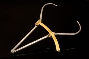 A photograph of a set of medical callipers set against a black background. The two arms of the device are made from a metal resembling steel or aluminium, and a curved brass ruler extends from the mid-point of the left arm, connecting to the right. The arms connect at the bottom of the device with a rivet and are straight until after the measuring tool, where each arm bows outward, curving in.
