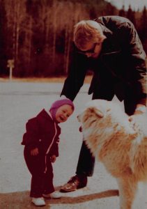 a faded, vintage photograph of a little kid in a red snowsuit and a pink and white winter hat. She wears white shoes. She is standing face-to-face with a fluffy white dog who has his tongue out. A man stands between the child and the dog, one hand on each, to make sure that the interaction remains friendly and safe. The man wears brown shoes, blue jeans, a dark jacket and sunglasses. His sandy blonde hair is shaggy. These figures stand on concrete and the sun casts shadows on the ground. In the background are trees and a sign that is blurry and out of focus.