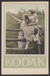 A hand-coloured photographic image of two white women in a rustic scene with the brand name Kodak printed underneath in large capital letters. Both women are facing left and are wearing short-sleeved gingham dresses, one of them is wearing a similarly patterned hat and is sitting on the fence holding an old box-style camera. The other woman stands behind the fence and points off to the left of the image. Behind the women are the rolling hills of the countryside with trees on the horizon line.