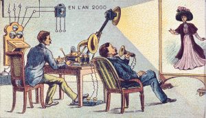 A colour lithograph illustration of two white men with brown hair and moustaches in late 19th century suits sitting in chairs next to a table. The man on the left is sitting directly behind the table and is operating a phonograph and a projection device that sits on the table, projecting an image of a white woman in a purple flowing dress and flowered hat on a screen to the right. The other man sits to the right of the table and speaks into a horn-shaped device. Against the wall are various other devices of unknown use. the words En L'An 2000 appear next to these devices in block lettering.