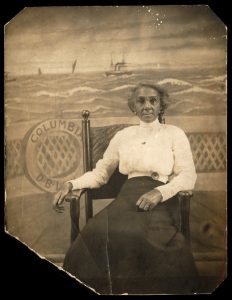 A photograph of a woman sitting on a chair against a painted backdrop of a nautical scene. The woman is wearing a long black skirt and a puffy white blouse. She is wearing round glasses and looks directly at the camera. the painted backdrop recreates the deck of a ship looking off towards open water in the distance with ships of various types traveling the across the horizon.