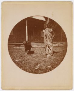 A sepia-toned circular photograph of a white man and a dark-coloured dog. The two figures are in front of a brick house, standing on grass littered with a few branches. The dog is looking off to the left and is in mid-step. The man is dressed in light-coloured trousers and a dark long-sleeve shirt and suspenders. The man's head is not in the frame, so we only see him from the shoulders down.
