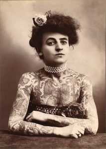 A black and white photograph of a woman sitting at a table. Her forearms are resting on the surface of the table, and she is covered in elaborate tattoos from the neck down. She has a multi-rowed pearl necklace and a flower in her hair.