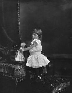 A black and white photograph of a young white girl playing with a doll in an interior space. The space is dark, with visible curtains, and decorative moulding in the background. The girl's body is turned to the left of the image with her head turned slightly towards the camera without looking directly at it. She is wearing a frilly white dress and is standing on a chair. She holds her doll with both arms, standing the toy on a table.