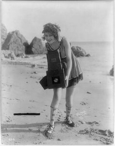 A black and white photographic image of a young woman standing on a sandy beach. The woman has a delighted expression on her face and is wearing a short dress with no sleeves, socks pulled up to her shins, and intricately laced shoes. She is leaning to the left, and holding a large box-style camera which is open, prepared to take a photograph.