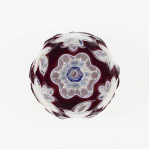 A photograph of a glass paperweight against a white background. The paperweight is a rough sphere, with recessed white floral patterns in a deep reddish-purple body. A central design contains an intricate decoration of white red, and various shades of blue.