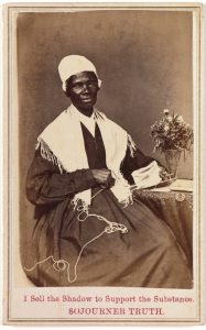 A sepia-toned photograph of an African-American woman sitting next to a small table that stands to her left. The woman is wearing a long-sleeved black dress, a white cloth head covering, and a white shawl covering her shoulders. She is holding fabric and knitting implements in her hands, with her left arm resting on the table. A vase of flowers sits on the table, which is covered in a dark, patterned cloth.