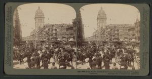 A sepia-tone stereoscopic image of a busy city street. A large number of people are moving in a variety of directions against a backdrop of towering buildings. The tallest building is centrally located and reaches to the very top of the image. The majority of the figures are wearing dark clothing, and are making their way through the streets amidst trolleys, horses and buggies. The scene is so populated, it is easy for the individual to get lost in the chaotic nature of the image.