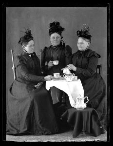 A black and white photograph of three white women sitting around a table having tea. The women are wearing long black dresses with matching ornate hats. A white cloth is draped over the table, on which the tea service sits.