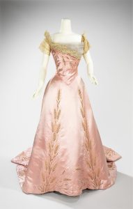 A colour photograph of a headless white mannequin wearing a short-sleeved pink ballgown. Yellow lace decorates the shoulders, with embroidered floral patterning through the dress.