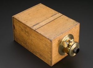 A colour photograph of a closed box-style camera from a three-quarter perspective. The camera's body is constructed from light-coloured wood fastened together with nails and dovetailing. There are two worn hinges on top of the box. At the front of the camera, the lens mechanism is prominently displayed. It is primarily constructed from brass and has a circular base that attaches to the wooden body. A brass cylinder extends from the base terminating in a smaller open-ended black cylinder. on top of the lens mechanism is a small dial that extends to the left via a thin rod.