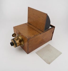 A colour photograph of an open box-style camera from a three-quarter perspective. The camera's body is constructed from light-coloured wood fastened together with nails and dovetailing. At the front of the camera, the lens mechanism is prominently displayed. It is primarily constructed from brass and has a circular base that attaches to the wooden body. A brass cylinder extends from the base terminating in a smaller open-ended black cylinder. on top of the lens mechanism is a small dial that extends to the left via a thin rod. The back end of the camera is open, with a section of wood pivoting from the near centre point to a 90-degree angle. Black light-resistant material is seen on the underside of this open section. A piece of square photographic paper lies to the front-right of the camera.