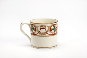 A photograph of a mug set against a white background. The mug is off-white with gold pinstriping along the base and rim. The pinstriping also borders decorative designs that span the top half of the mug. The decoration is a leaf design, incorporating gold, red and green.