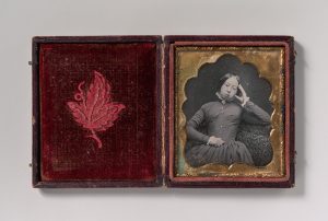 A photographic image of an open photo case. The case opens like a book revealing two panels. The left panel is lined with a worn red fabric, embossed with a leaf. The right panel contains a black-and-white photograph of a white woman framed in a decorative gold frame. The woman is sitting on a chair, her head resting slightly against her left hand. The woman is wearing a long sleeved dark dress with white lace accents on her collar and wrists.