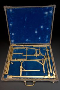 An open case lined with blue fabric. The bottom portion of the case contains a number of brass measuring tools of varying descriptions. They are neatly laid out, consisting mostly of right angles, save for two curved implements.