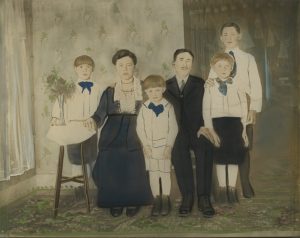 A photographic image of a six-member white family, located inside of a home, that is hand-coloured. The mother and father are wearing dark clothing and are sitting in chairs. The children are wearing white tops with blue ribbons tied around their necks.
