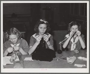 A black and white photograph of three young white women knitting white seated at a long table. The women are each concentrating on their work with fabrics and yarn of varying light and dark shades.