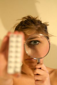 A photographic image of a white woman holding a blurry out-of-focus pill box in her right hand towards the camera. In her left hand is a magnifying glass held up to her left eye.