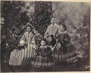 A photograph of an outdoor scene. A white woman and four white girls are grouped centrally in the image, surrounded by various bushes. A large tree covered in ivy rises behind the figures. These figures are wearing long dresses, three of which appear to be identical dark dresses, with the rightmost child holding a hat in her hands. The woman is seated and wears a vertically striped light dress and holds a hat in her right hand resting on her lap. A child wearing a lighter dress stands at the back of the group holding a hat in her hands.