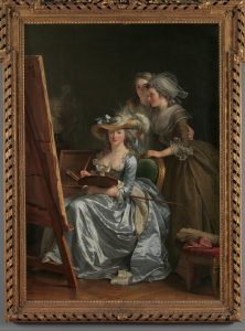 A framed oil painting of three white women. One of the women is sitting on an upholstered chair, holding a palette and brushes, her body turned toward a large easel and canvas to the left. She is wearing a lavish long-sleeved blue dress with lacing around the neckline. Atop her intricately styled grey-brown hair is an ornate hat decorated with feathers. The other two women stand behind her, looking at the canvas. These two women are wearing long dresses adorned with lace, one brown and one white.