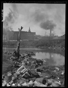 A black and white photograph of an industrial scene surrounding a small centrally-located body of water. Numerous factories appear in the background, all made from brick and producing thick black smoke from their respective smokestacks. An array of refuse is built up along the shores of the water, and a dead tree stands to the left.