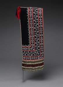 A photograph of an ornately beaded and decorated hood positioned that only a rectangular patch of cloth is seen. Bands of decorations from long vertical bands of various designs, turning a right angle at the bottom. The designs are all on a black background with red borders and white patterning in between. Strings of beads hand from the bottom of the fabric, all having the same colour pattern.