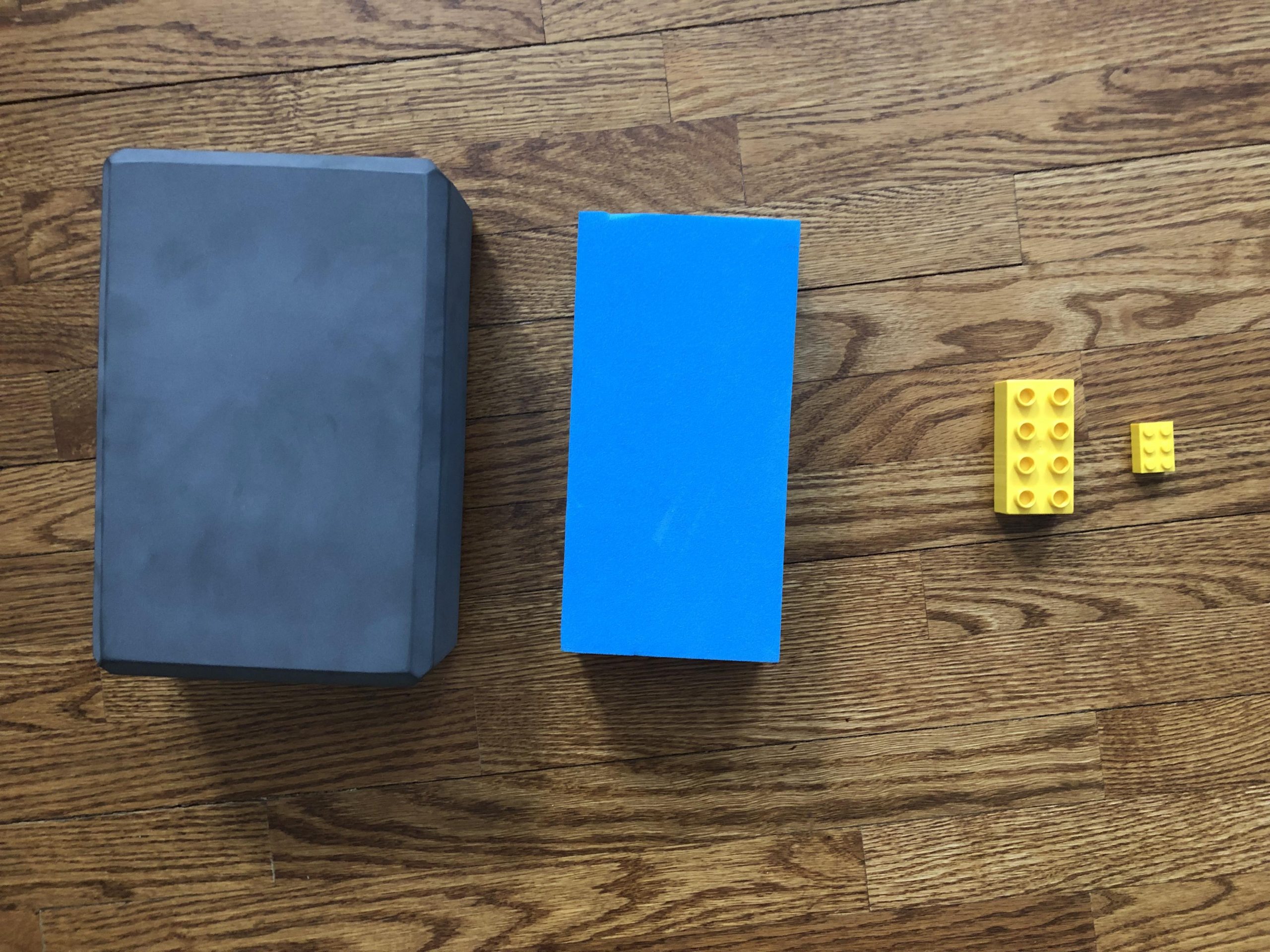 Photo of four blocks on a wooden floor. Two are large foam blocks, a grey one slightly larger than a blue one. Two are small yellow Lego blocks, one smaller than the other.