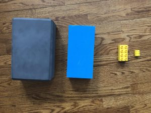 Photo de quatre blocs sur un plancher en bois. Deux sont de gros blocs en mousse, un bloc gris légèrement plus gros qu’un bloc bleu. Deux sont de petits blocs jaunes, l’un plus petit que l’autre.