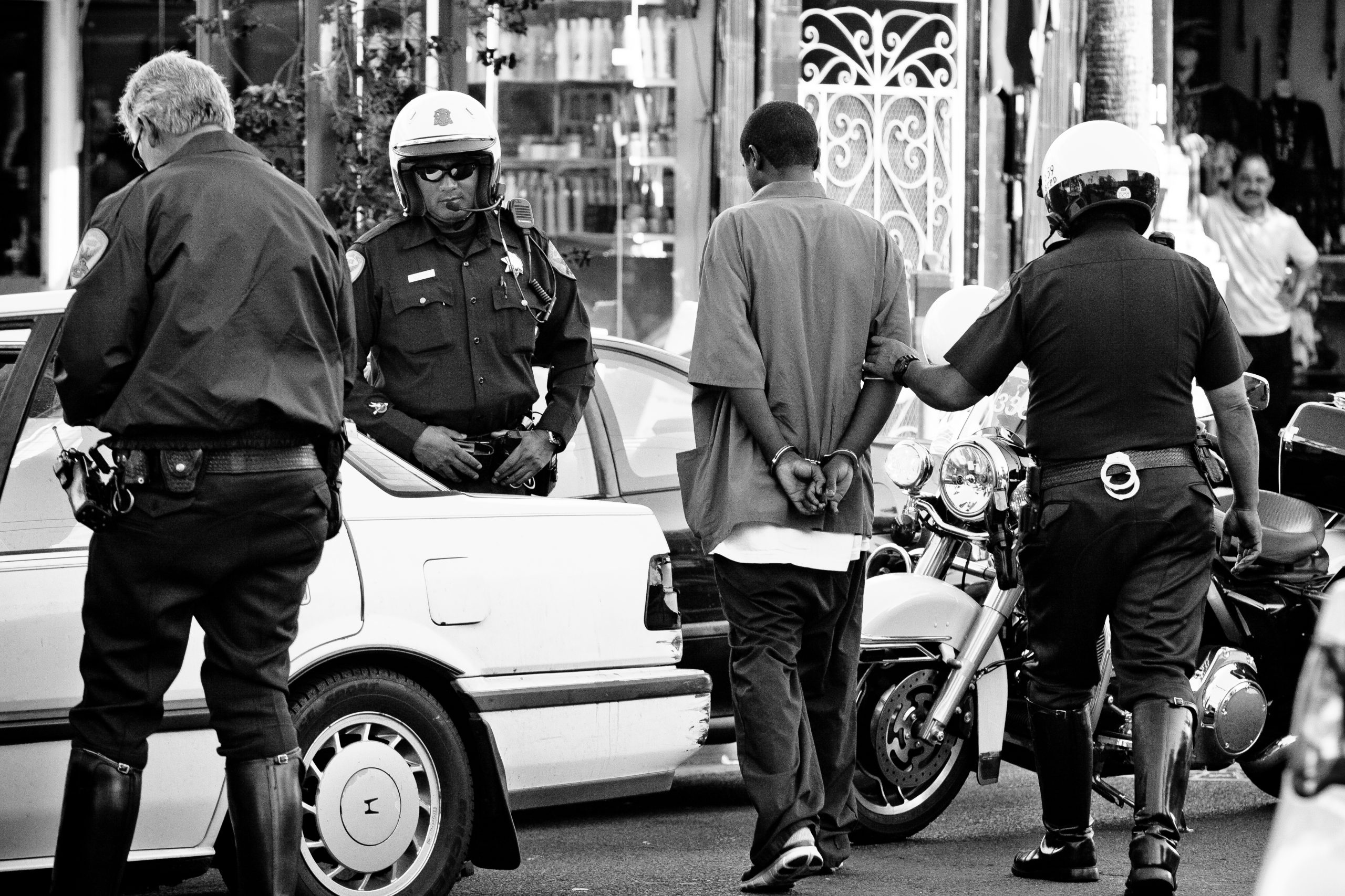 Photo of a 3 police officers, including 2 wearing motorcycle helmets, walking a young black man handcuffed behind his back.