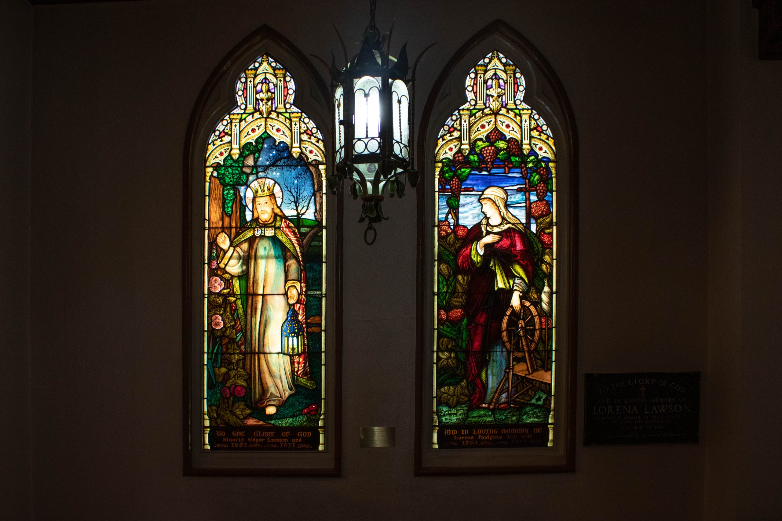 Two stained glass windows. Left window shows man knocking at a door while holding a lantern. The right windows shows a woman touch a spinning wheel.
