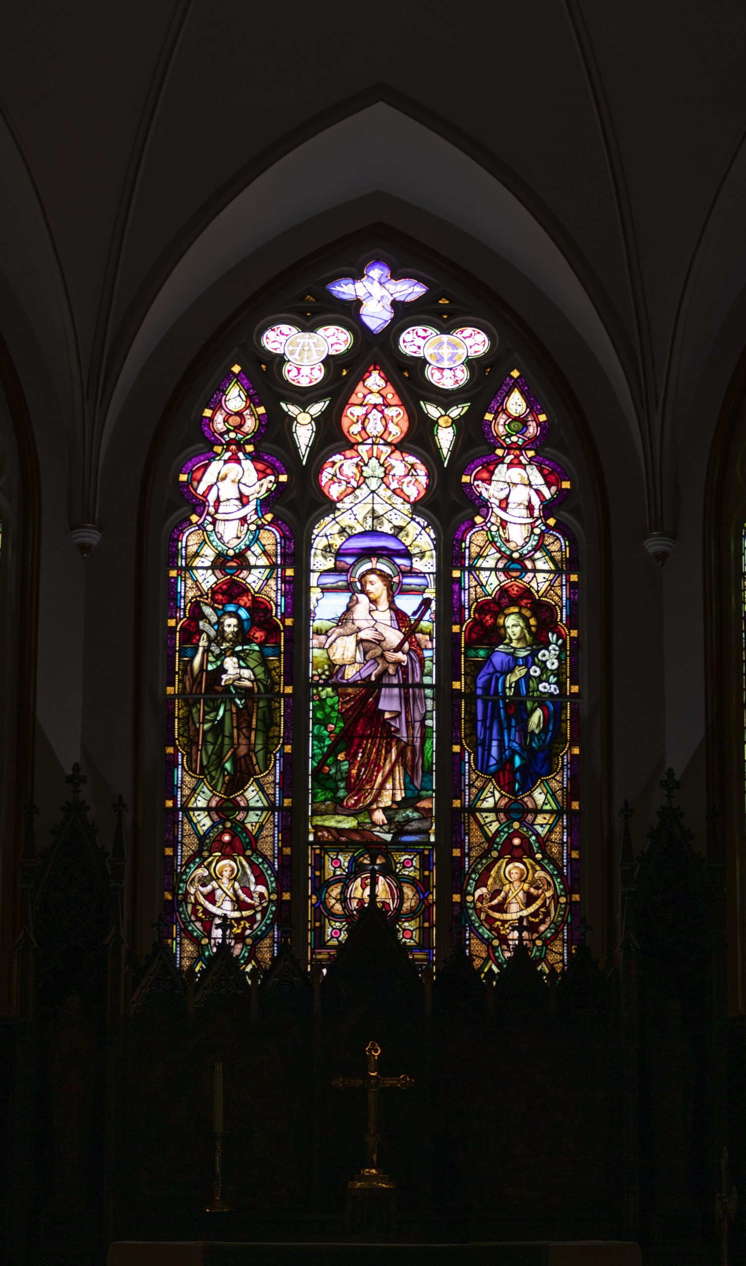Three stained glass windows. Center image depicts a man holding a lamb.